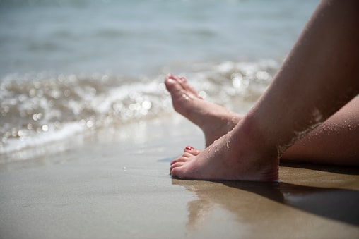 Bare feet at the water's edge on a sandy beach, illustrating the comfortable, pain-free movement CarePlus helps hammertoe patients in Bellevue keep.